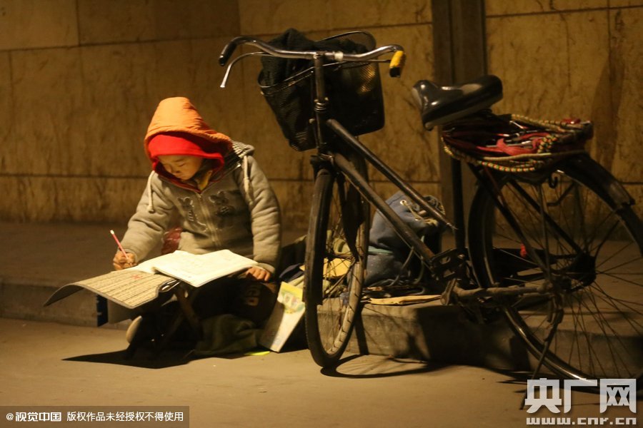 鄭州中原路京廣鐵路大橋下涵洞的馬路牙子邊，一位戴著紅色辣椒帽的小男孩趴在一個小馬扎上，上面鋪著一張車墊，在微弱的燈光下寫作業(yè)